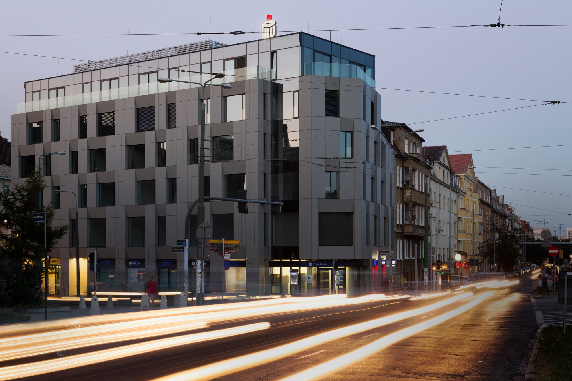 Nowoczesny budynek usługowy z szarą elewacją, widoczny z perspektywy ulicy z rozmazanymi światłami przejeżdżających samochodów, na dachu logo firmy.