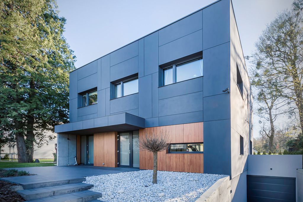 Nowoczesny dom z szarą elewacją z paneli, drewnianymi akcentami i minimalistycznym ogrodem z białych kamieni oraz małym drzewkiem.