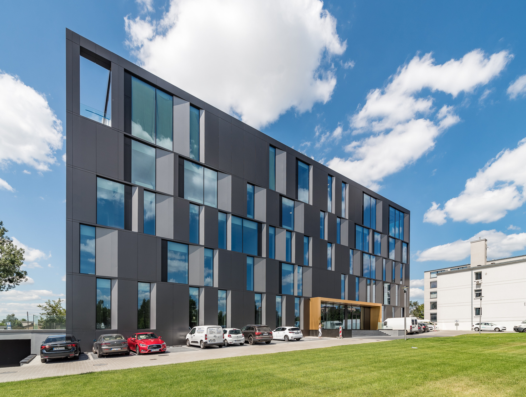 Nowoczesny budynek biurowy z ciemną elewacją, dużymi oknami odbijającymi niebo, trawnikiem i zaparkowanymi samochodami przed wejściem.