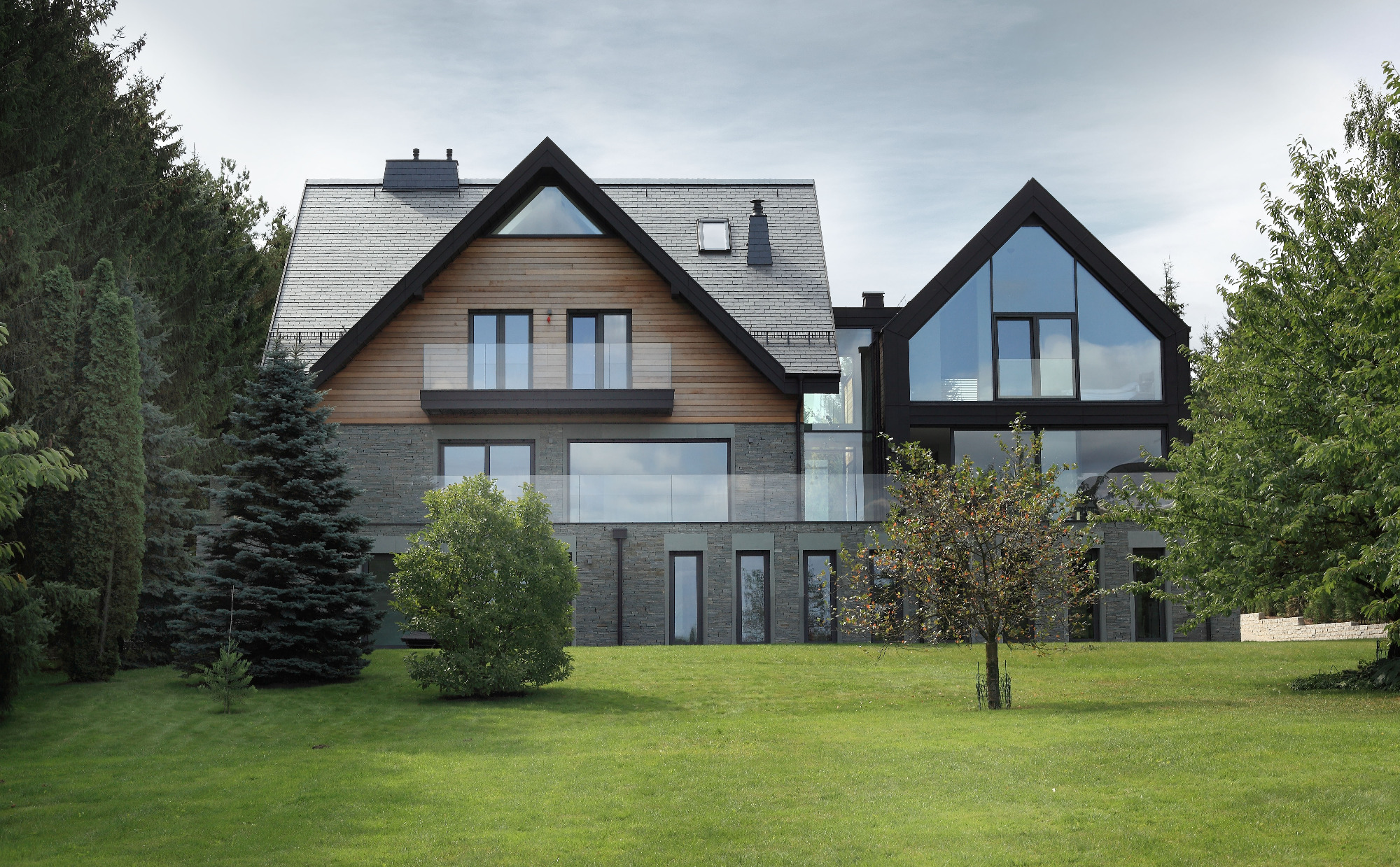 Nowoczesny dom jednorodzinny z dwuspadowym dachem, elewacją z kamienia i drewna, dużymi przeszkleniami i zielonym trawnikiem na pierwszym planie.