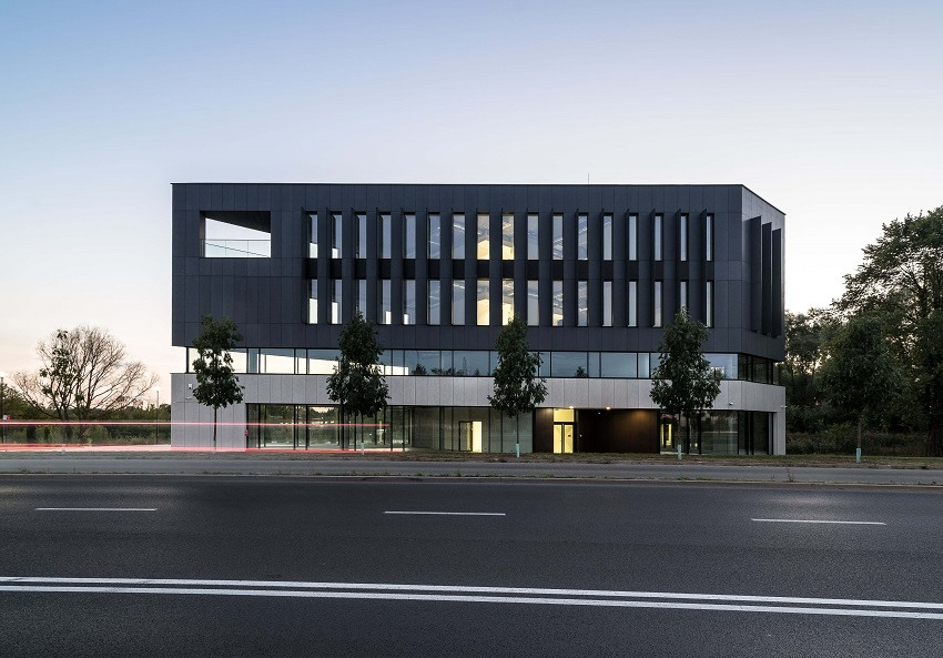 Nowoczesny budynek biurowy z ciemną elewacją i pionowymi oknami, widok z poziomu ulicy z rozmazanymi światłami przejeżdżających samochodów.