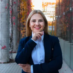 Portret uśmiechniętej kobiety w granatowym żakiecie i białej koszuli, z ręką podpartą pod brodą, na tle kolumn porośniętych jesienną roślinnością.