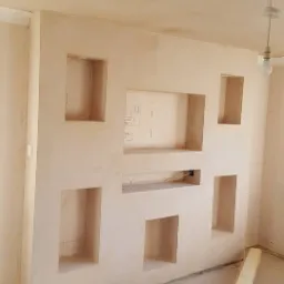 Ściana z wnękami po szpachlowaniu, widoczne ślady ołówka, surowy betonowy sufit, podłoga z narzędziami i materiałami budowlanymi.