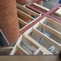 Wnętrze w trakcie budowy, widok z góry przez okno, ukazujący drewniany szkielet konstrukcji stropu z czerwonymi stalowymi belkami i fragmentem murowanego komina z cegły.