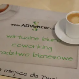 Filżanka espresso na podstawce z logo firmy oferującej doradztwo biznesowe, coworking i adres korespondencyjny, obok leży długopis.