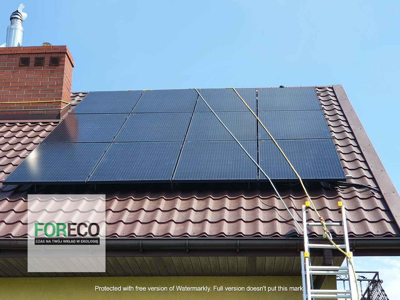 Panele fotowoltaiczne na brązowym dachu z dachówki ceramicznej, zabezpieczone linami, drabina oparta o rynnę, komin z cegły w tle, błękitne niebo.