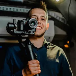 Uśmiechnięty mężczyzna z brodą i irokezem pozuje z profesjonalną kamerą Sony na stabilizatorze, trzymając go w dłoni. Widoczne oświetlenie studyjne i fragment sufitu w tle.