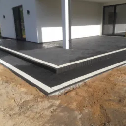 Nowoczesny taras z ciemnej kostki brukowej obrzeżonej jasnym granitem, prowadzący do domu z dużymi przesuwnymi drzwiami tarasowymi, widoczne przygotowane podłoże z piasku.