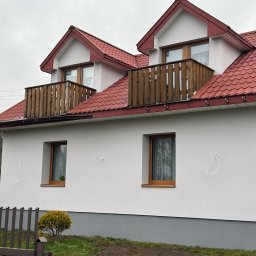 TermoBud Rafał Łochowski - Dwukondygnacyjny dom z czerwonym dachem i drewnianymi balkonami, świeżo otynkowany na biało, z widocznymi śladami po montażu na elewacji.