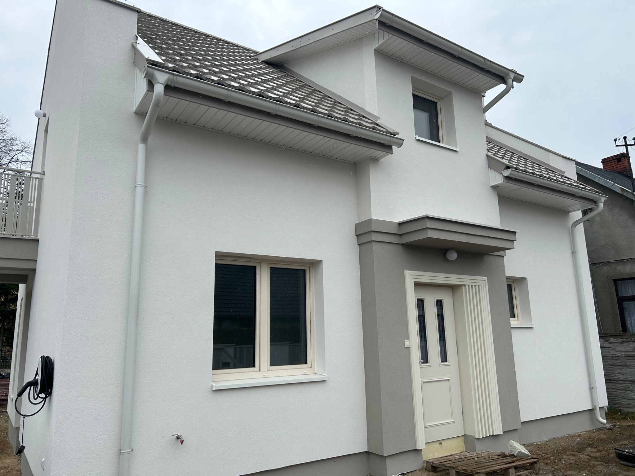 Biały dom z szarą stolarką okienną i drzwiami wejściowymi, widok z zewnątrz. Nowoczesna elewacja z widocznymi rynnami i fragmentem balkonu.