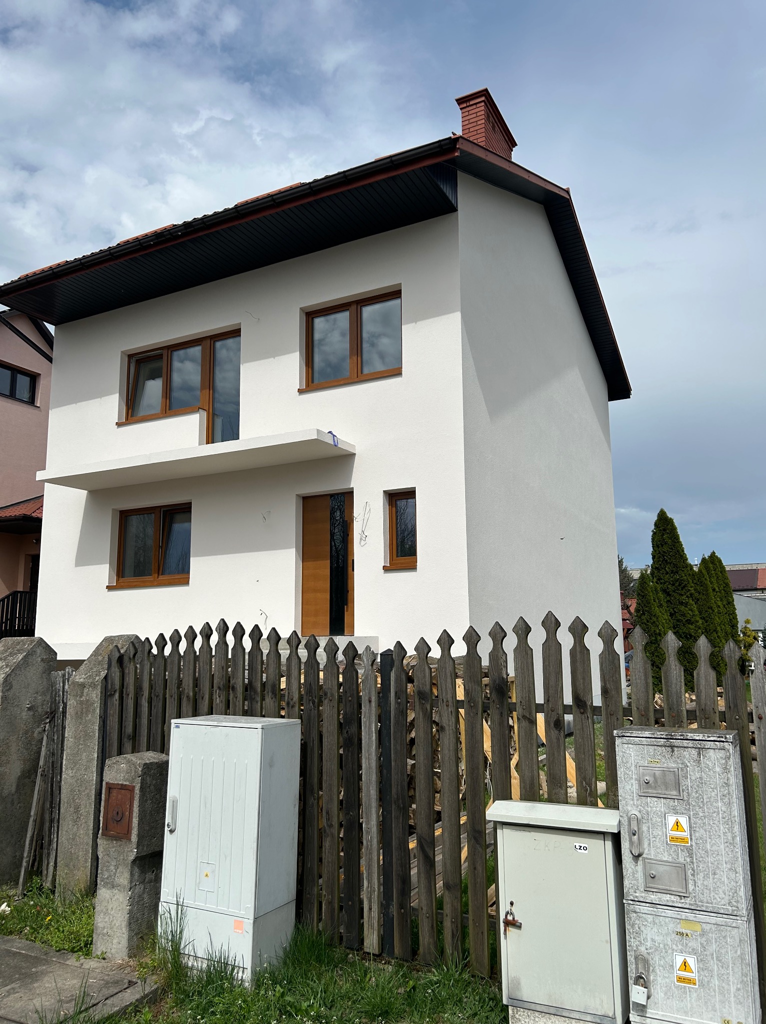 Biały, dwukondygnacyjny dom z brązowymi oknami i drzwiami, widziany zza drewnianego płotu. Widoczne skrzynki elektryczne i fragmenty sąsiednich budynków.