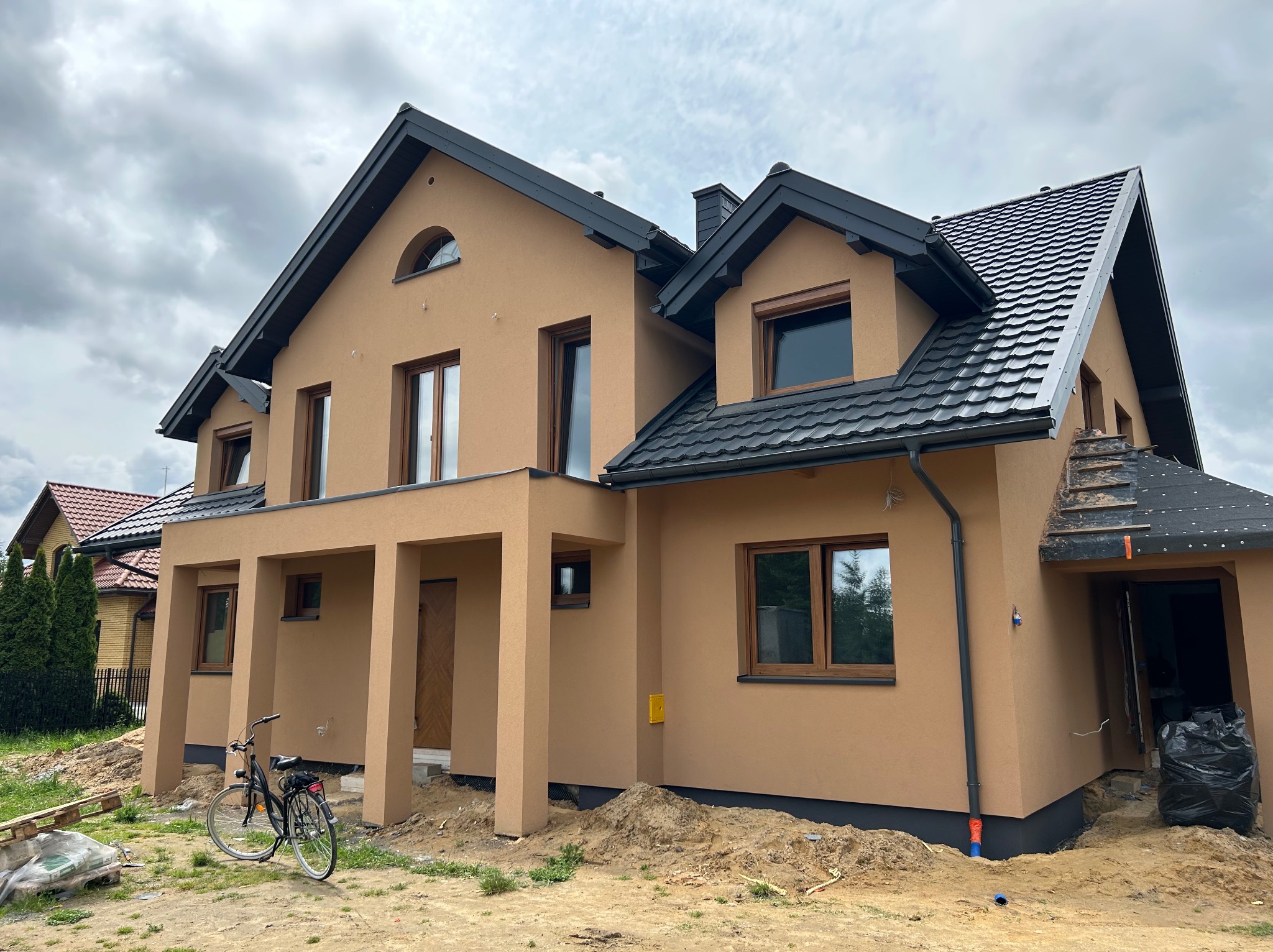 Nowoczesny dom jednorodzinny w Pionkach z elewacją w odcieniach beżu i brązu, czarną dachówką i rowerem opartym o kolumnę. Widok z zewnątrz, pochmurne niebo.