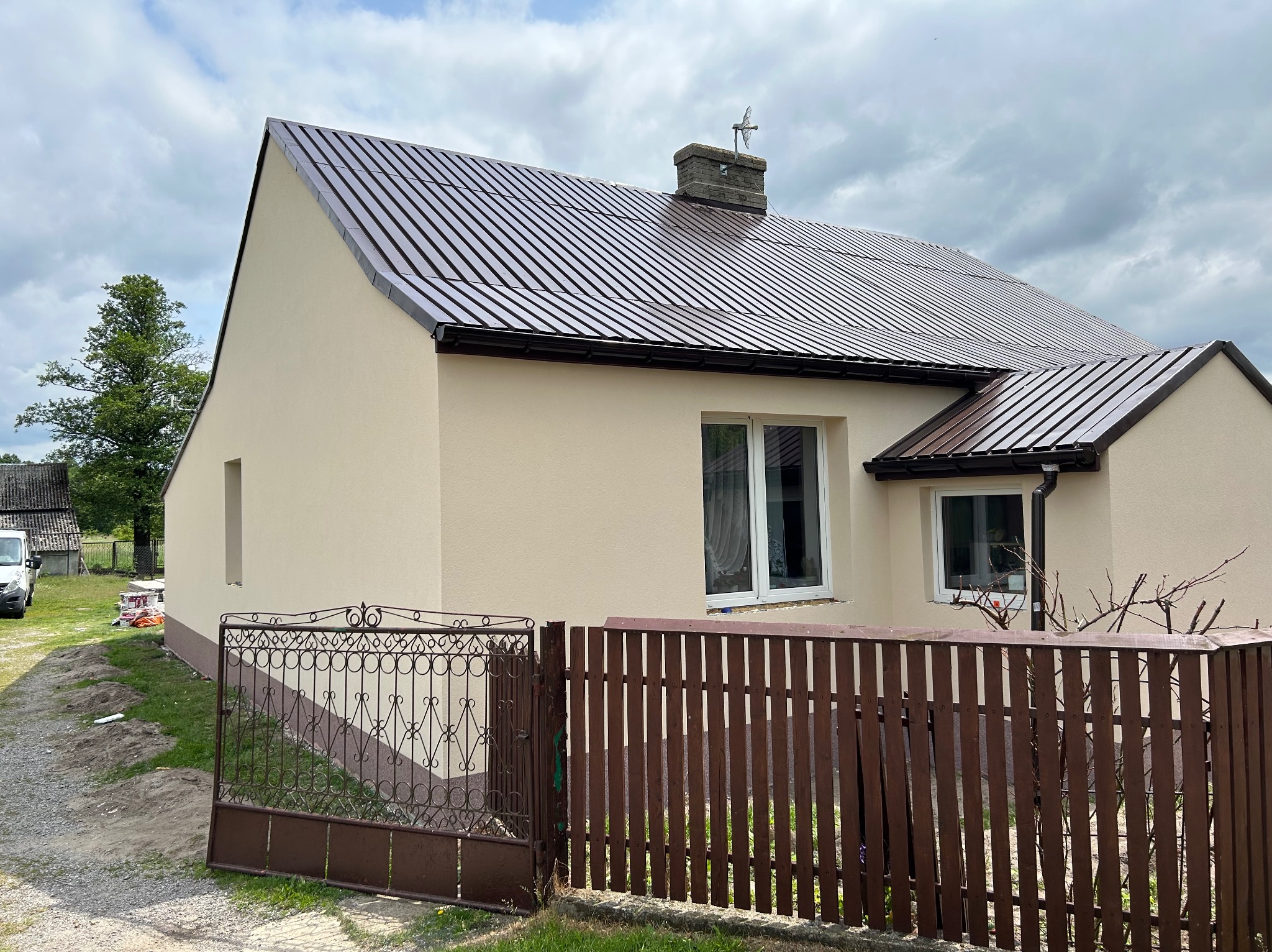 Dom jednorodzinny z beżową elewacją i brązowym dachem z blachy trapezowej, ogrodzony brązowym płotem. Widoczny komin i antena.
