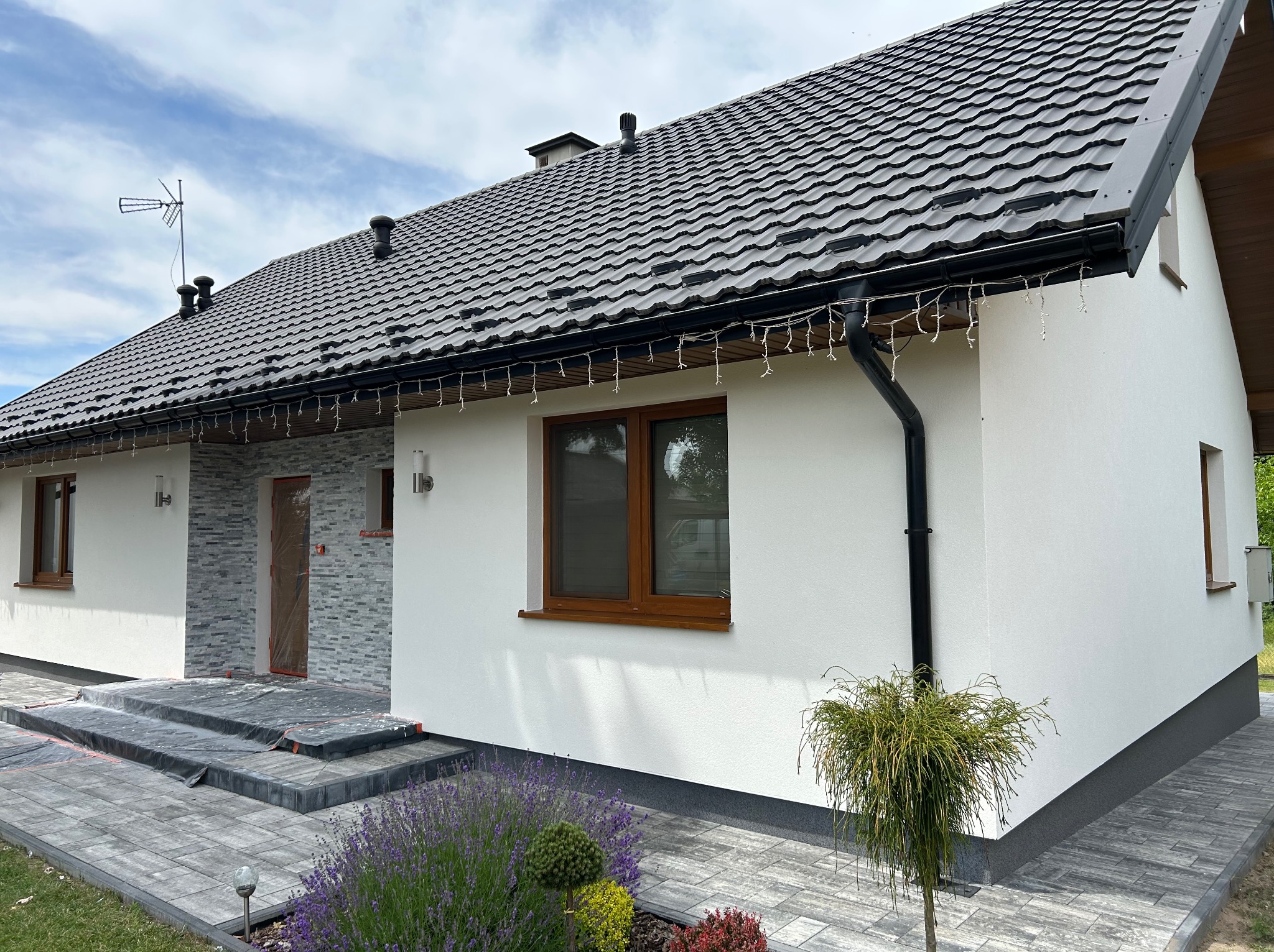 Biała elewacja domu jednorodzinnego z ciemną dachówką i kamienną okładziną przy wejściu. Widoczne elementy dekoracyjne i zadbany ogród.