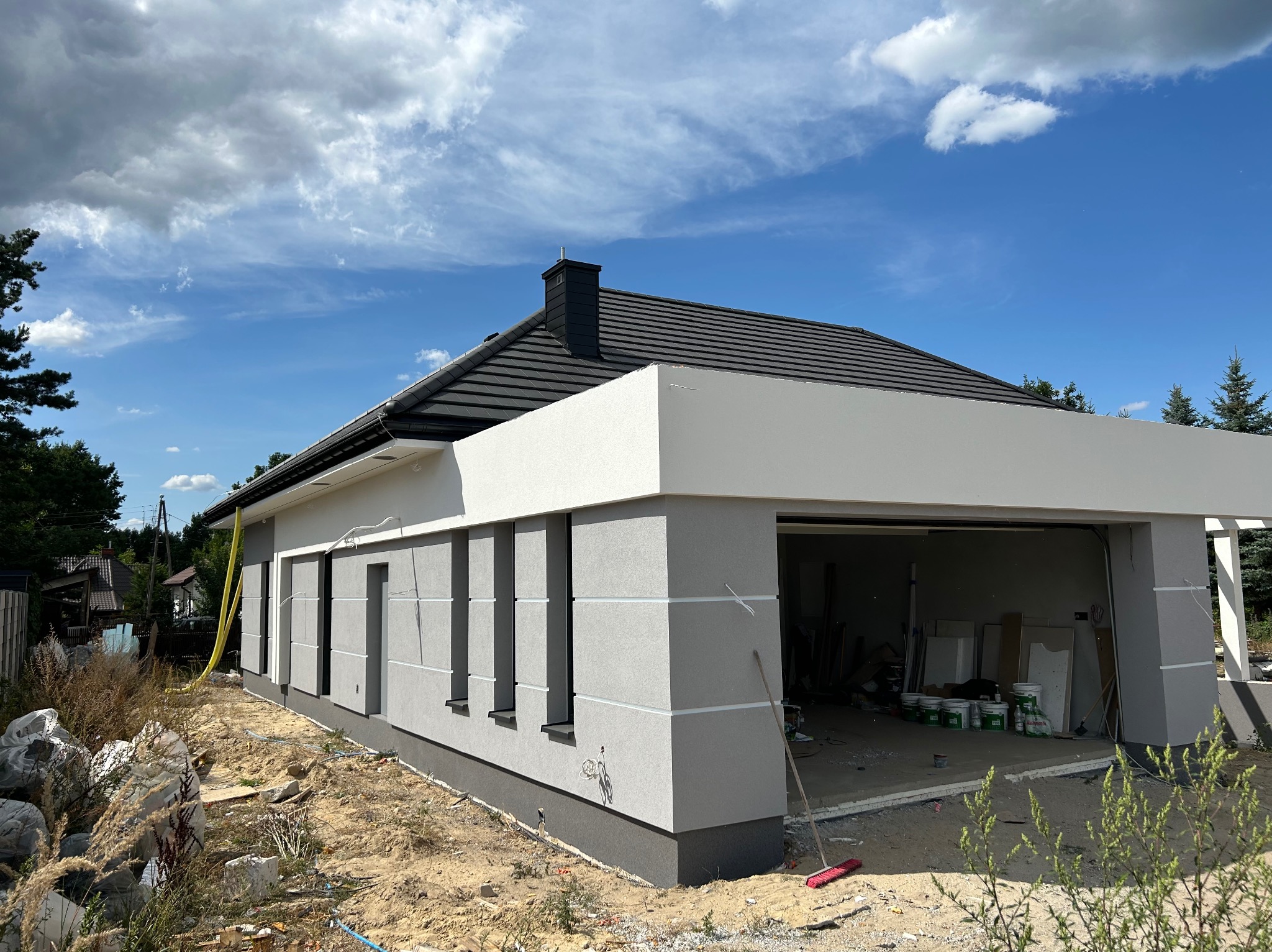 Nowoczesny dom jednorodzinny z garażem w trakcie wykończenia elewacji. Widoczne szare ściany z białymi elementami dekoracyjnymi i czarny dach. Na podwórku farby i narzędzia.