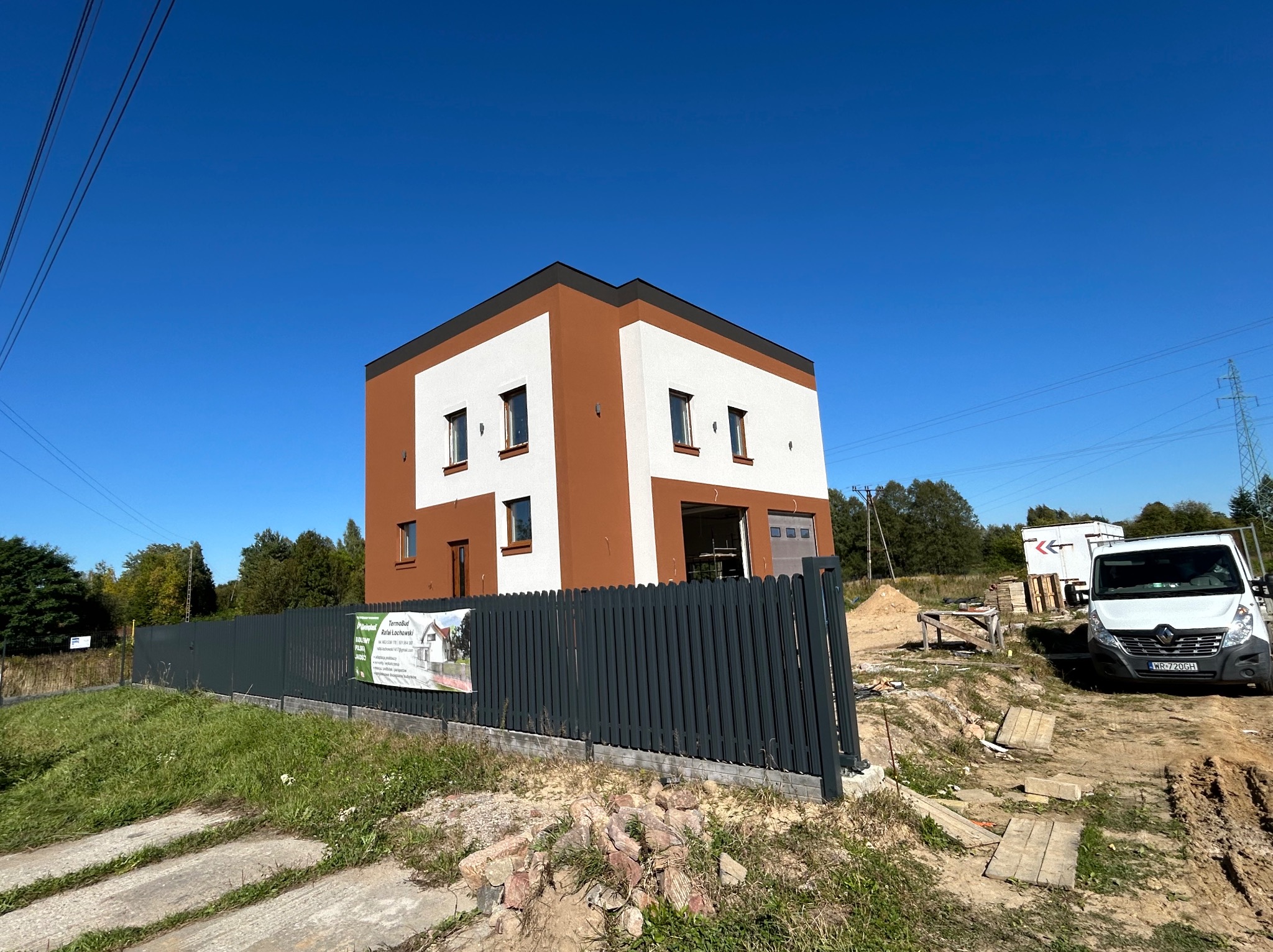 Dwukondygnacyjny dom z elewacją w kolorach bieli i brązu, ogrodzony szarym płotem. Na posesji zaparkowany biały bus. Błękitne niebo w tle.