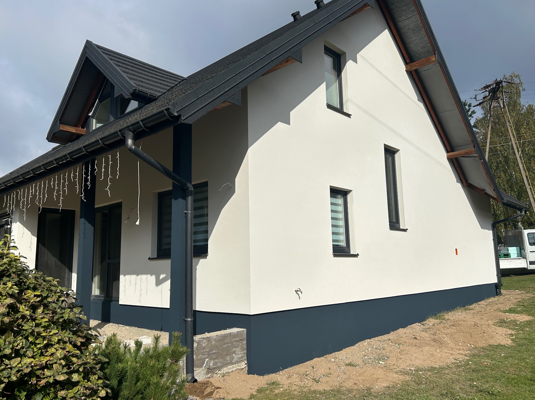 Nowoczesny dom jednorodzinny po renowacji elewacji, z białymi ścianami i ciemnoszarą podmurówką oraz stolarką okienną. Widoczne oświetlenie zewnętrzne.
