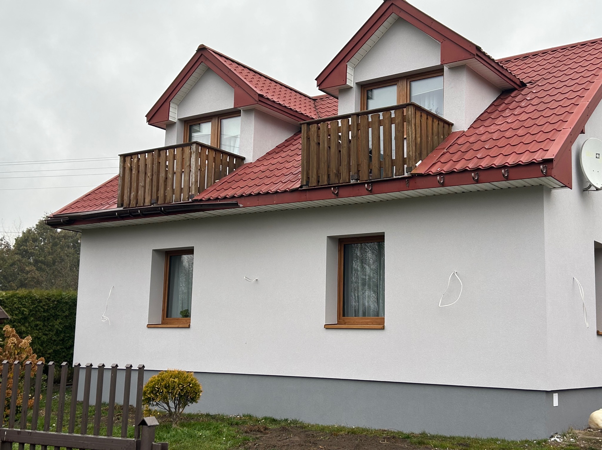 Dwukondygnacyjny dom z czerwonym dachem i drewnianymi balkonami, świeżo otynkowany na biało, z widocznymi śladami po montażu na elewacji.