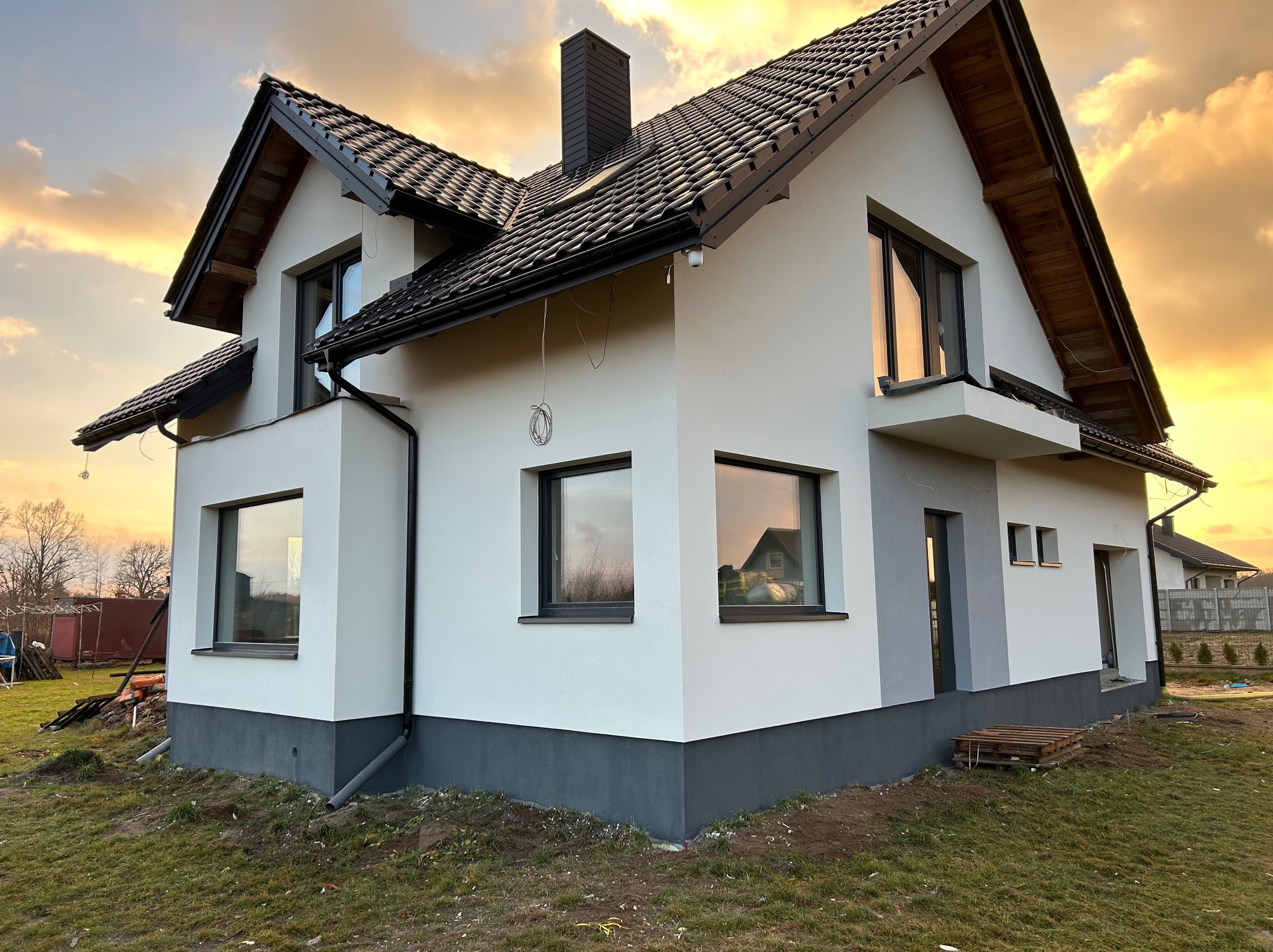 Nowoczesny dom jednorodzinny z białą elewacją i ciemną podmurówką, widoczny o zmierzchu. Ciemna dachówka i okna. Na trawniku widoczne palety.