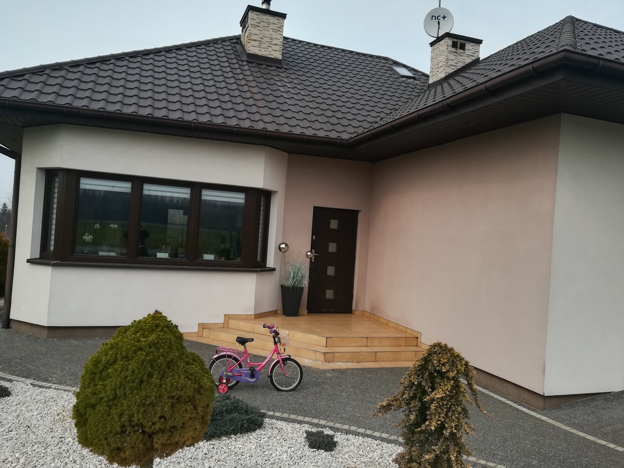 Dom jednorodzinny z jasną elewacją, brązowym dachem z dachówki, schodami wejściowymi wyłożonymi płytkami i różowym rowerem dziecięcym zaparkowanym przed wejściem.