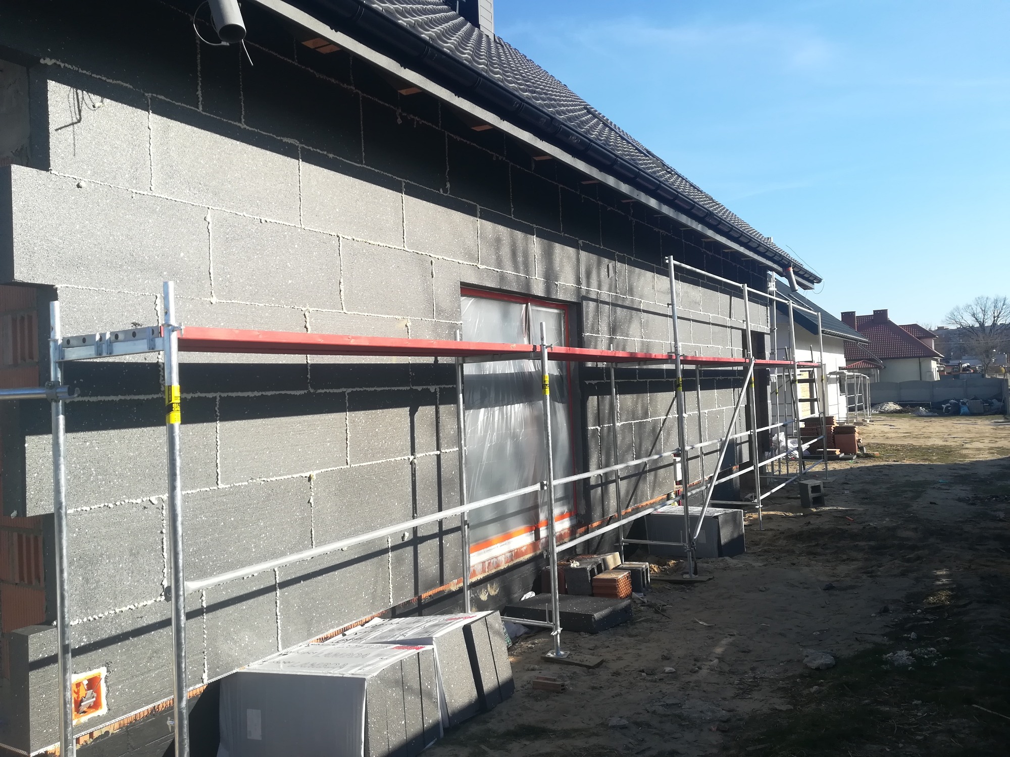 Dom jednorodzinny w trakcie ocieplania ścian zewnętrznych szarymi płytami izolacyjnymi, widoczne rusztowanie i materiały budowlane na posesji.