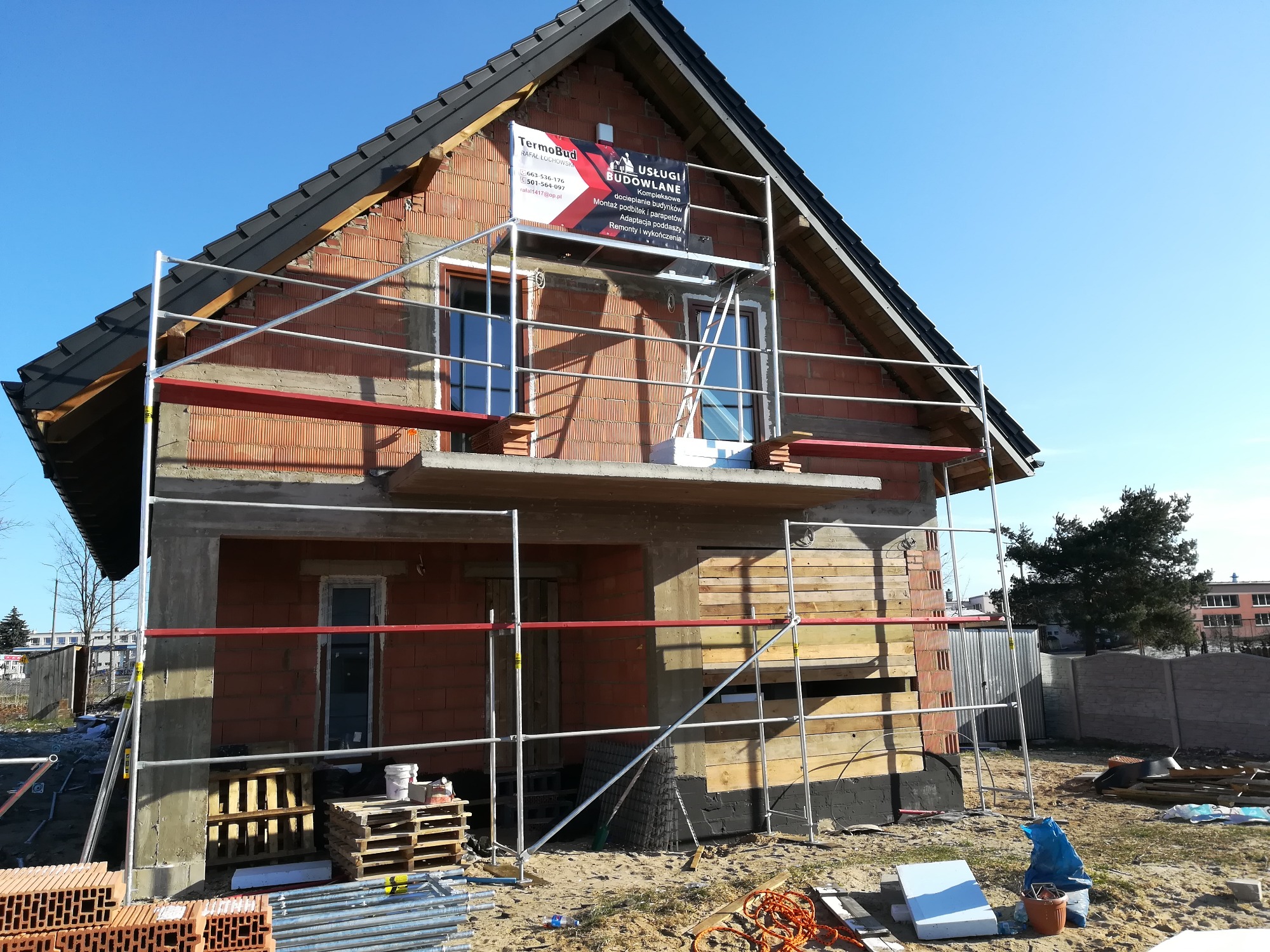 Dom w trakcie ocieplania elewacji, widoczne rusztowania, częściowo ocieplona ściana, cegły klinkierowe i baner reklamowy firmy budowlanej.