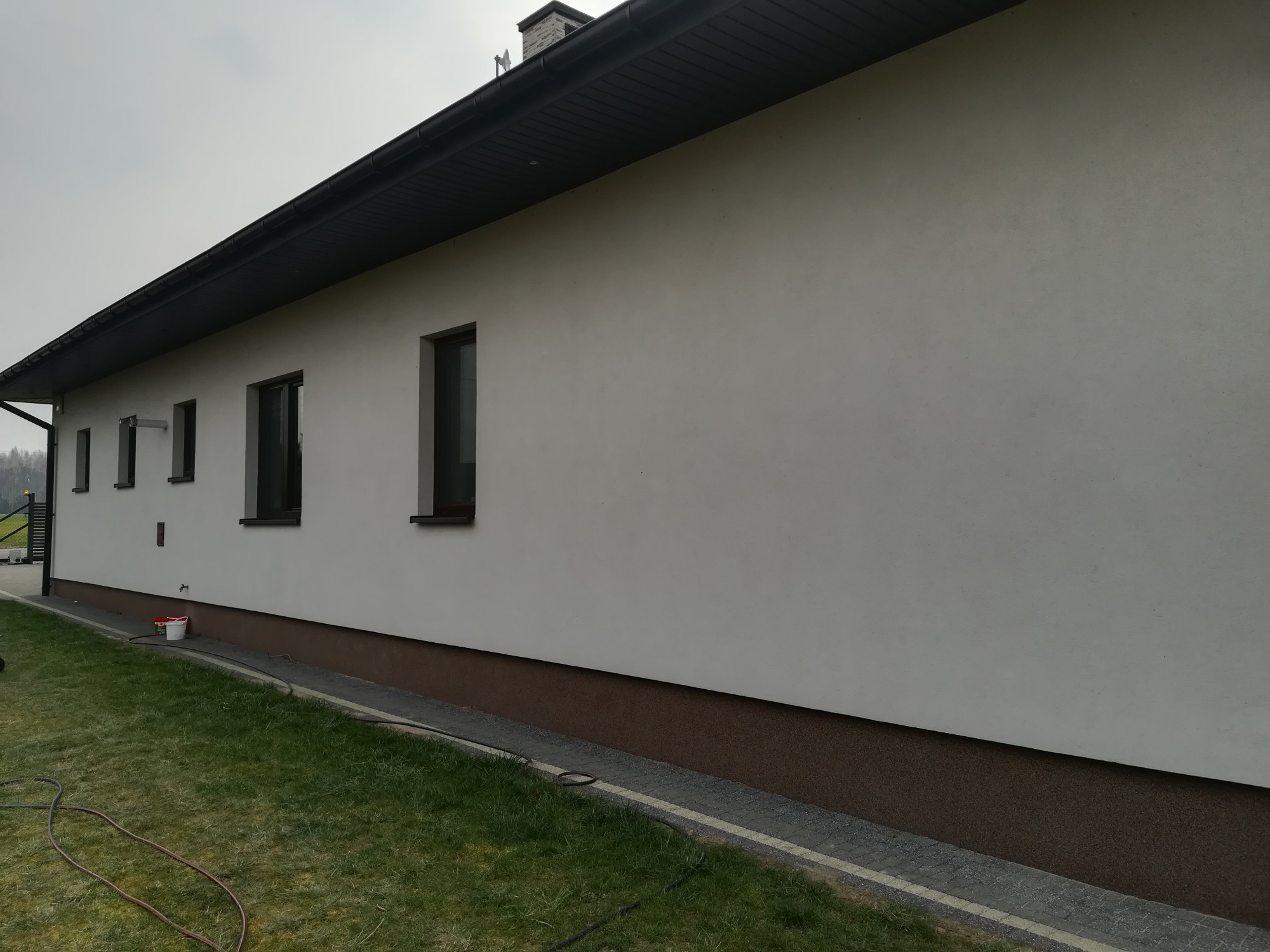 Świeżo otynkowana, gładka, biała elewacja domu jednorodzinnego z ciemnymi oknami i podbitką, widoczny fragment trawnika i wiadro z narzędziami przy ścianie.