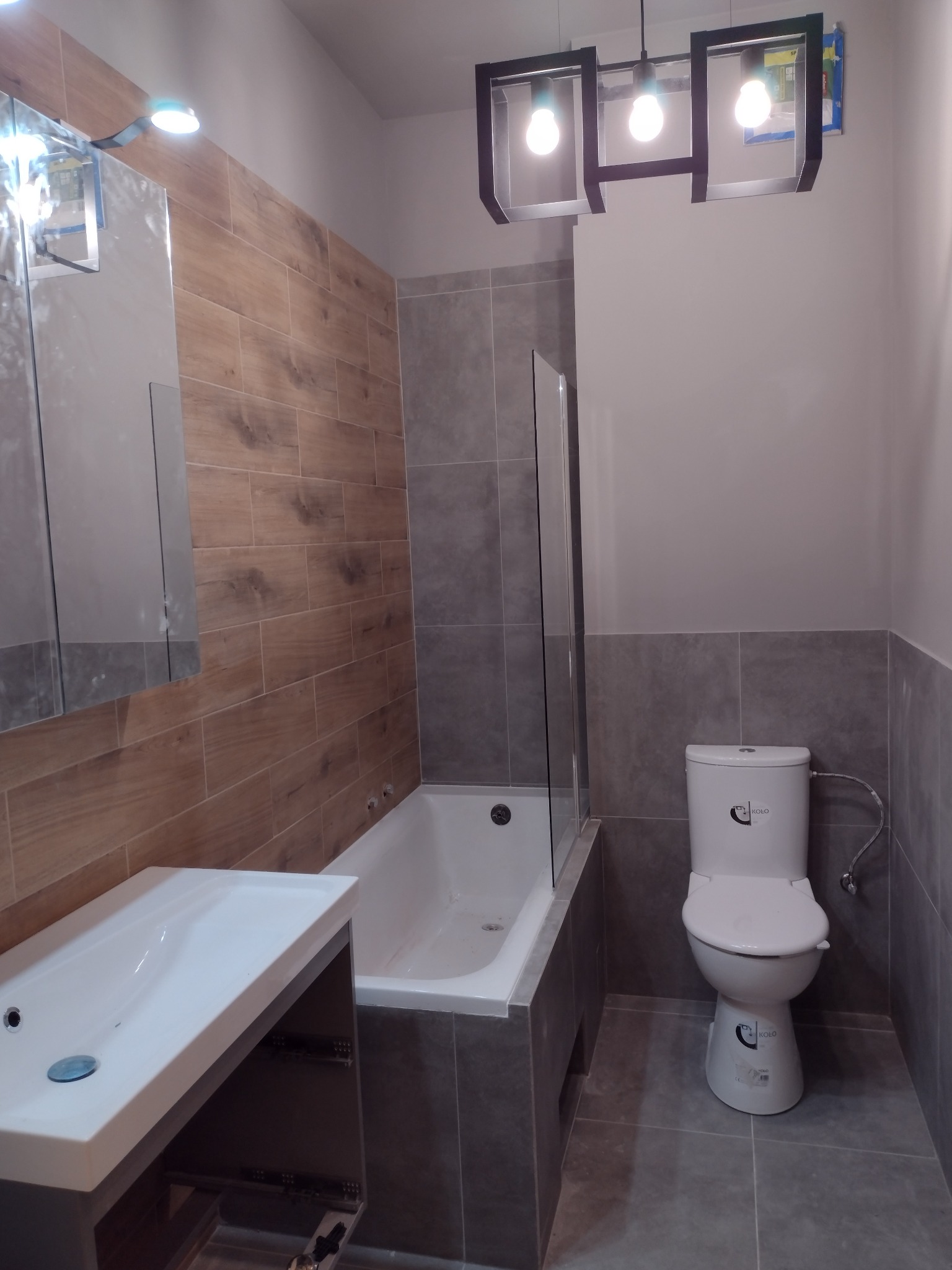 Wykończona łazienka z wanną zabudowaną szarą glazurą, umywalką z szafką w kolorze szarym, toaletą marki Koło i nowoczesną lampą wiszącą z trzema żarówkami.