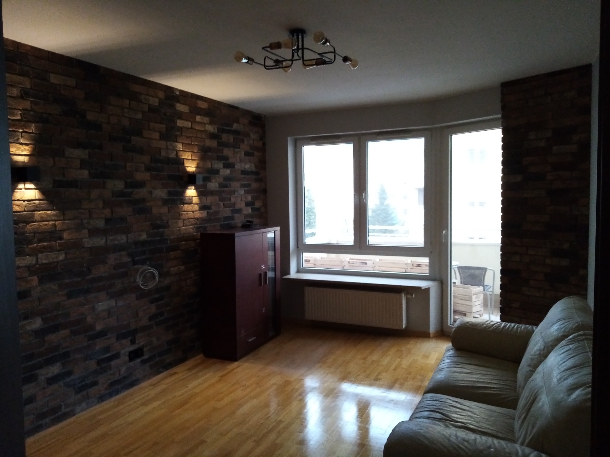 Pokój z ceglaną ścianą oświetloną kinkietami, parkietem i sofą, widok na balkon.