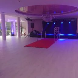 Sala bankietowa z czerwonym dywanem prowadzącym do sceny oświetlonej niebieskim światłem, stoły z nakryciami i krzesłami po lewej stronie, kolumny i żyrandol na suficie.