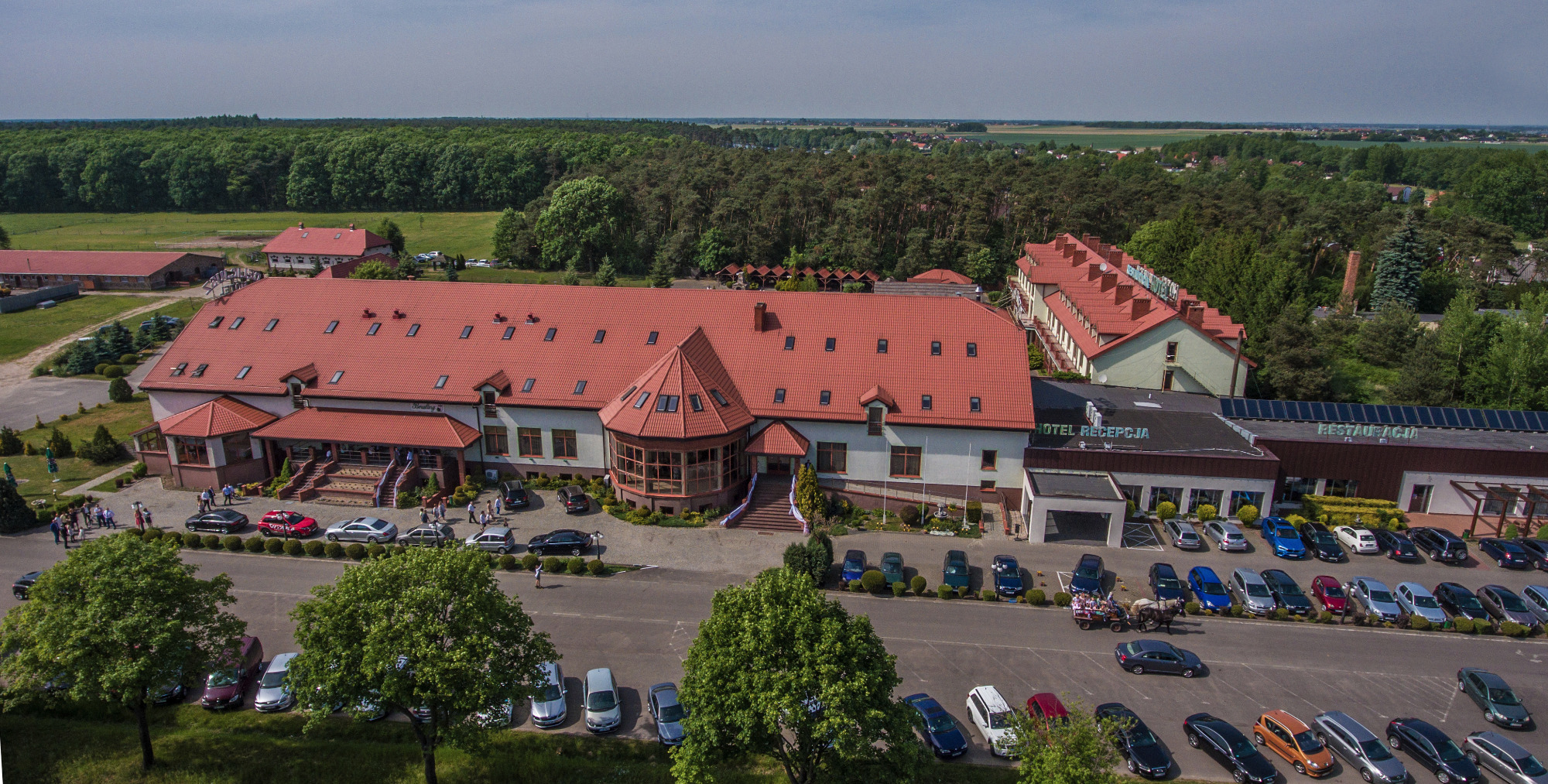 Panoramiczny widok na duży budynek hotelu z czerwonym dachem i parkingiem pełnym samochodów, w tle las i pola, bryczka konna na parkingu.