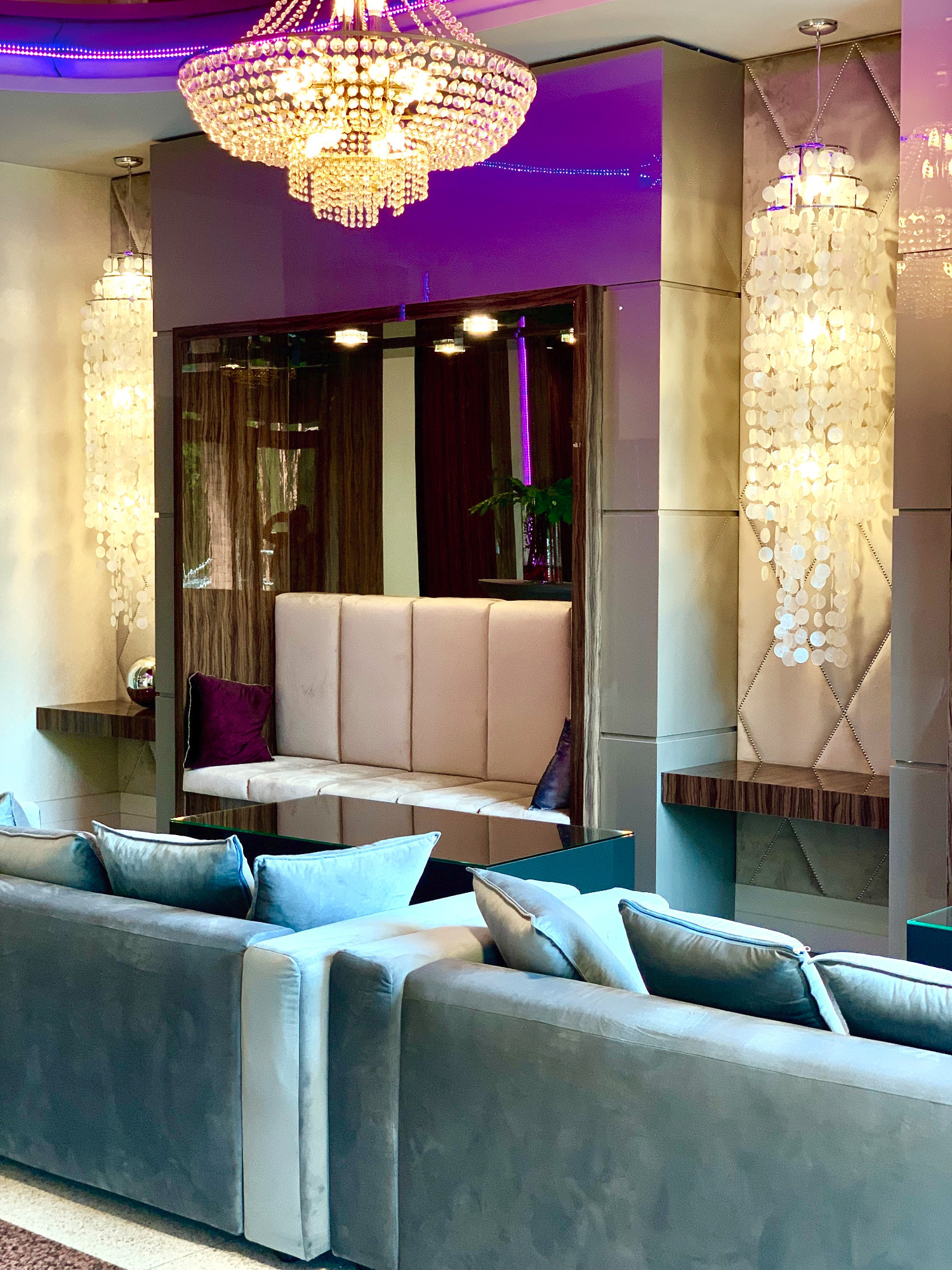 Eleganckie wnętrze hotelowe z szarymi sofami, dekoracyjnymi lampami z muszelek, kryształowym żyrandolem i fioletowym oświetleniem akcentującym