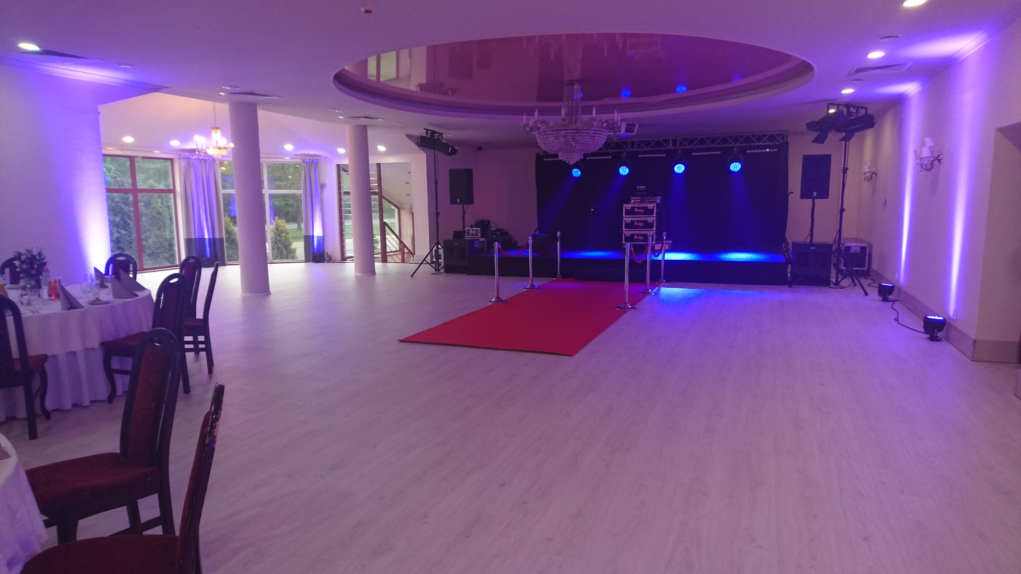 Sala bankietowa z czerwonym dywanem prowadzącym do sceny oświetlonej niebieskim światłem, stoły z nakryciami i krzesłami po lewej stronie, kolumny i żyrandol na suficie.