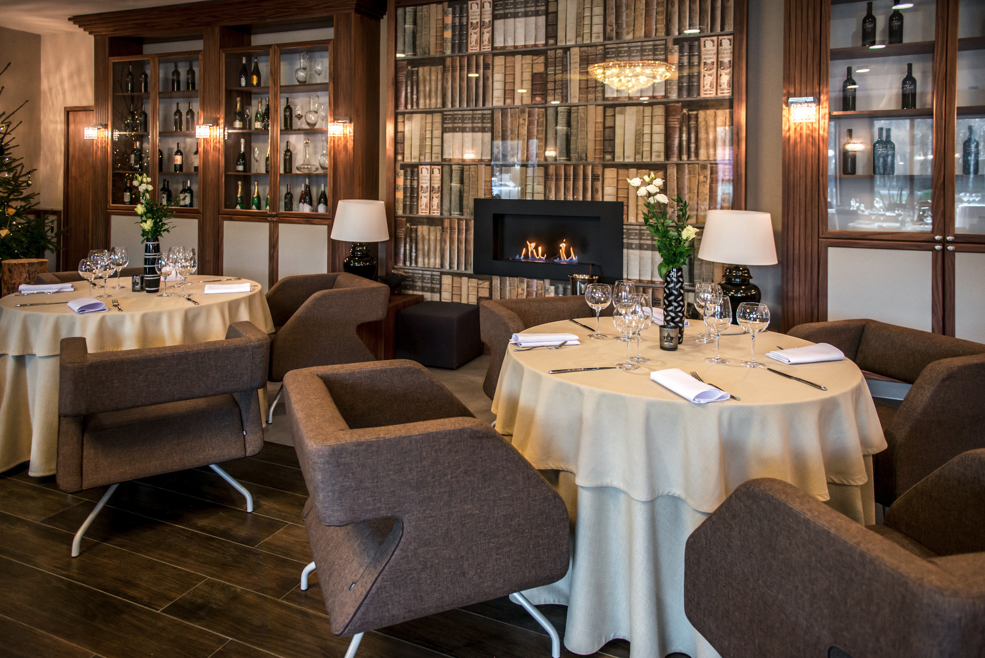 Elegancka sala restauracyjna z nakrytymi stolami, brązowymi fotelami i dekoracyjnym kominkiem na tle ściany z motywem biblioteczki, oświetlona lampami i żyrandolem.