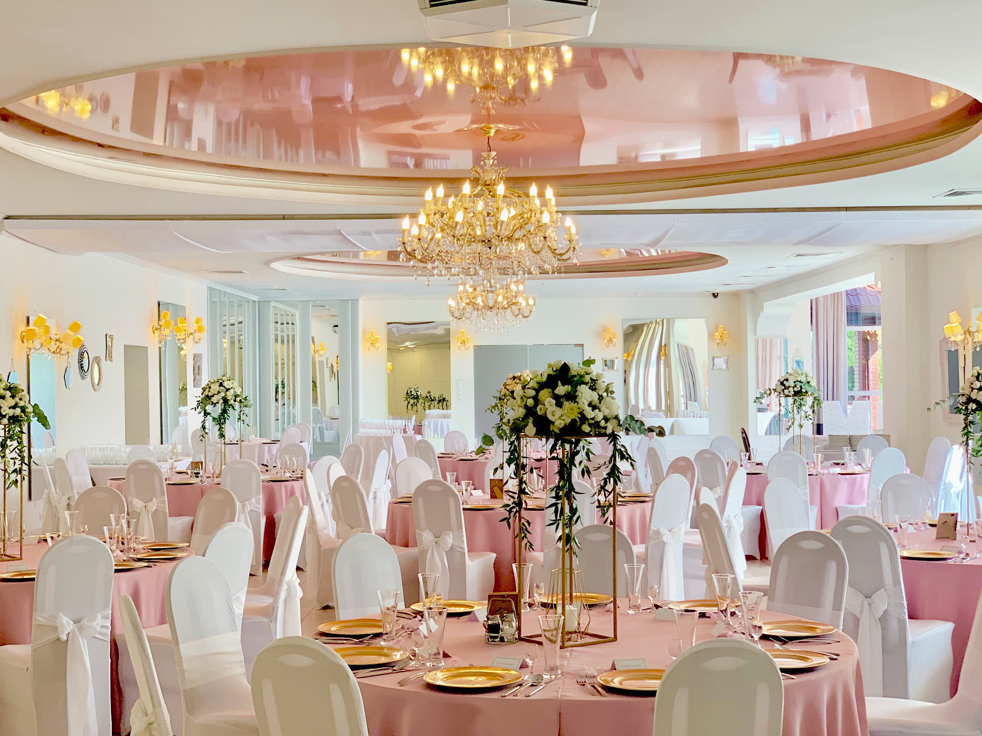 Sala bankietowa z okrągłymi stołami nakrytymi różowymi obrusami, białymi krzesłami z kokardami, złotymi talerzami i wysokimi kompozycjami kwiatowymi z zielonymi pnączami, oświetlona kryształowym...