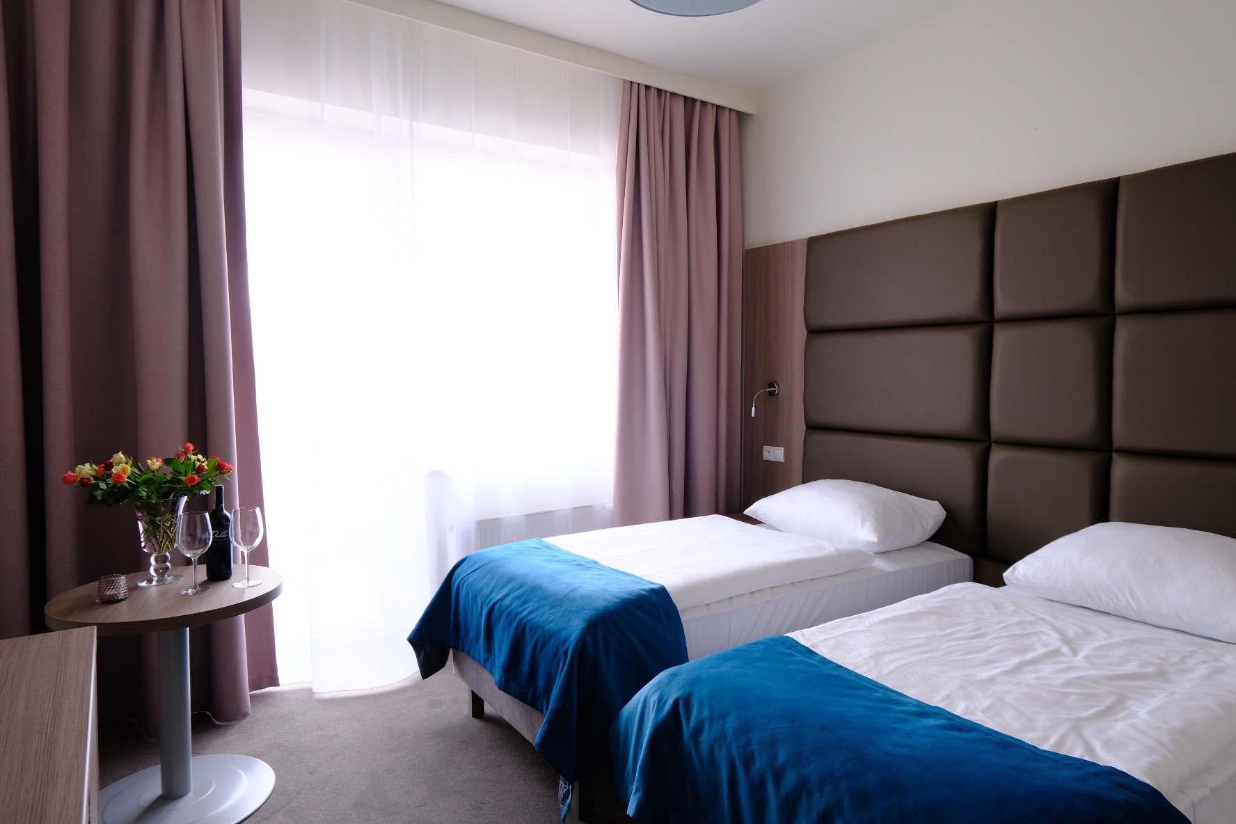 Pokój hotelowy z dwoma łóżkami pojedynczymi, nakrytymi białymi pościelami i niebieskimi narzutami, stolik z bukietem kwiatów, butelką wina i dwoma kieliszkami przy oknie z jasnymi zasłonami.