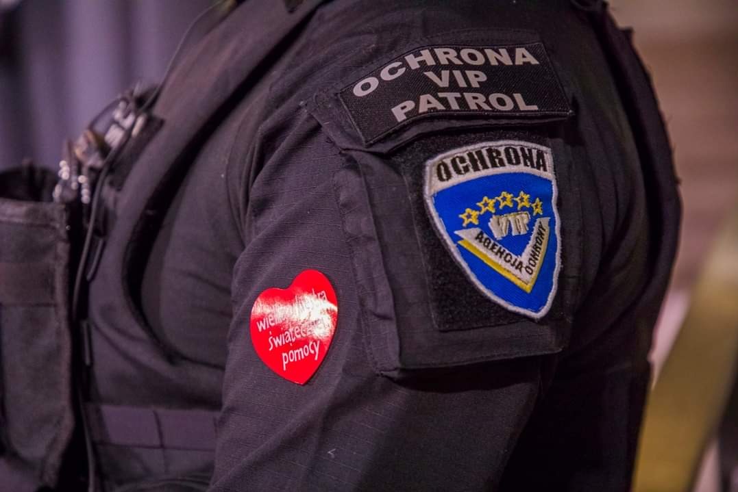 Czarny mundur pracownika ochrony z naszywkami 'OCHRONA VIP PATROL' i logo agencji ochrony, na rękawie czerwone serduszko 'Wielu Świątecznej Pomocy'.
