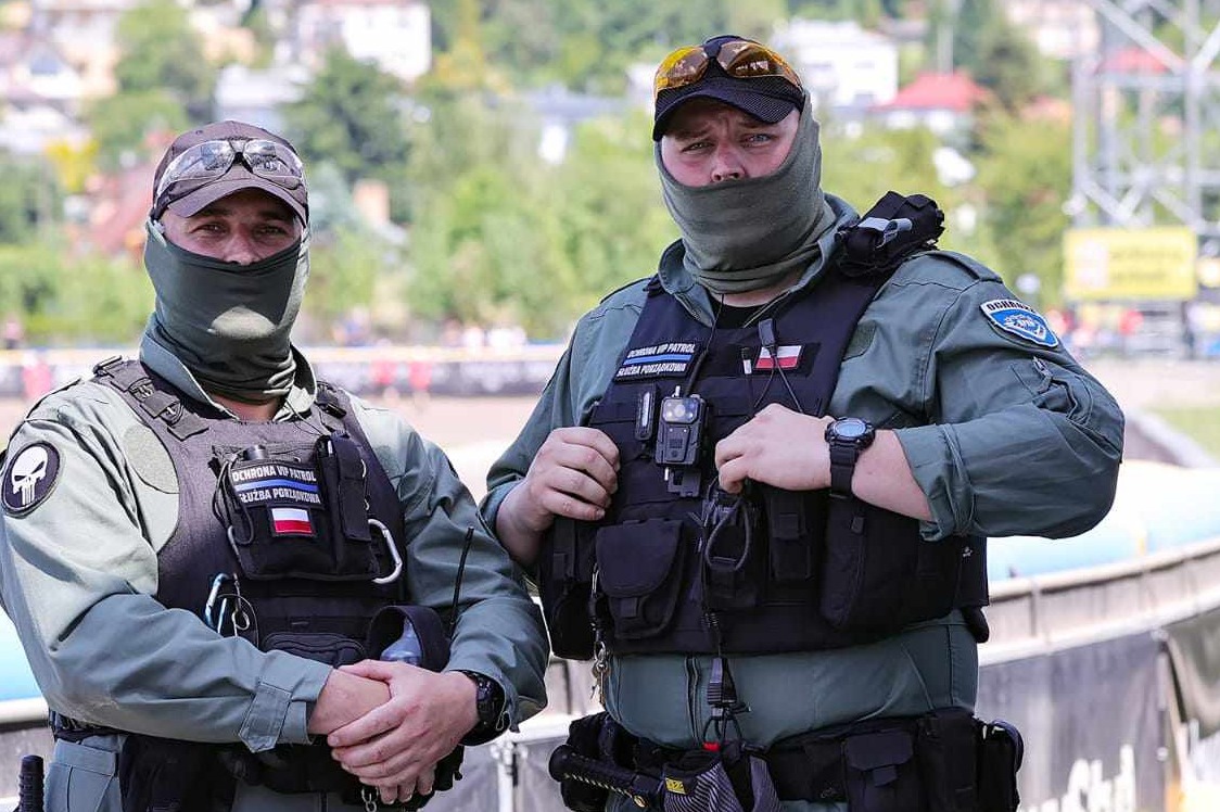 Dwóch umundurowanych mężczyzn w kamizelkach taktycznych i kominiarkach, z naszywkami 'Ochrona VIP Patrol' i flagą Polski, stojących na tle zieleni i trybun.