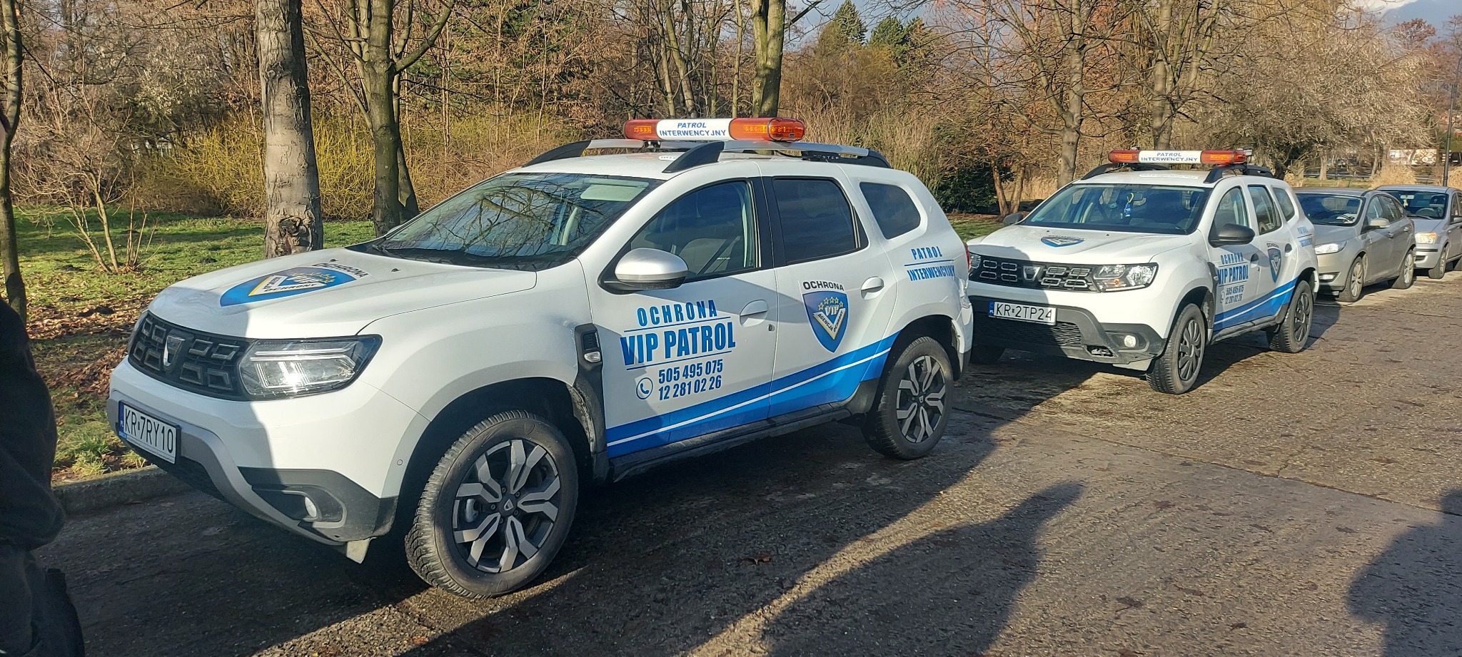 Dwa białe samochody patrolowe z niebieskimi akcentami i napisem 'OCHRONA VIP PATROL' zaparkowane obok siebie na tle parku z drzewami bez liści.