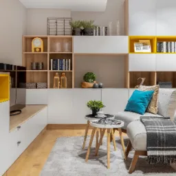 Jasne wnętrze pokoju dziennego z zabudową meblową: białe szafki z drewnianymi i żółtymi akcentami, drewniane półki z dekoracjami, szary dywan i fotel z poduszkami.