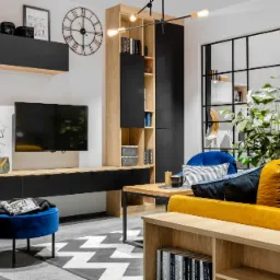 Nowoczesny salon z drewnianymi meblami w odcieniach beżu i czerni, z żółtą sofą i niebieskim pufem, uzupełniony geometrycznym dywanem i roślinami doniczkowymi.