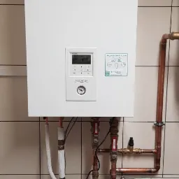 Biała pompa ciepła Panasonic Aquarea Smart Cloud zamontowana na ścianie z beżowych płytek, widoczne miedziane rury instalacji grzewczej z zaworami i sterownikiem.