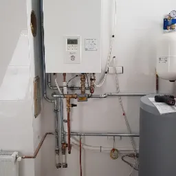 Biały piec grzewczy Panasonic z widocznymi rurami miedzianymi i elementami instalacji w pomieszczeniu z białymi płytkami, obok szary zbiornik i narzędzie na górze.