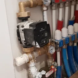 Rozdzielacz ogrzewania podłogowego z pompą obiegową Grundfos, manometrami i rurami PEX w kolorach niebieskim i czerwonym, zamontowany przy białej ścianie i brązowej podłodze.