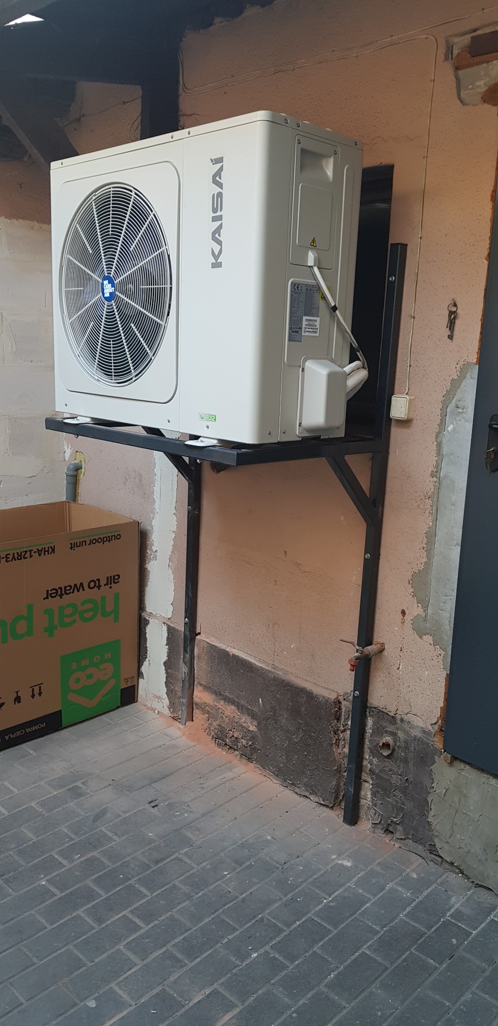 Zewnętrzna jednostka pompy ciepła Kaisai zamontowana na metalowym stelażu przy ścianie budynku z widoczną skrzynką kartonową po lewej stronie.