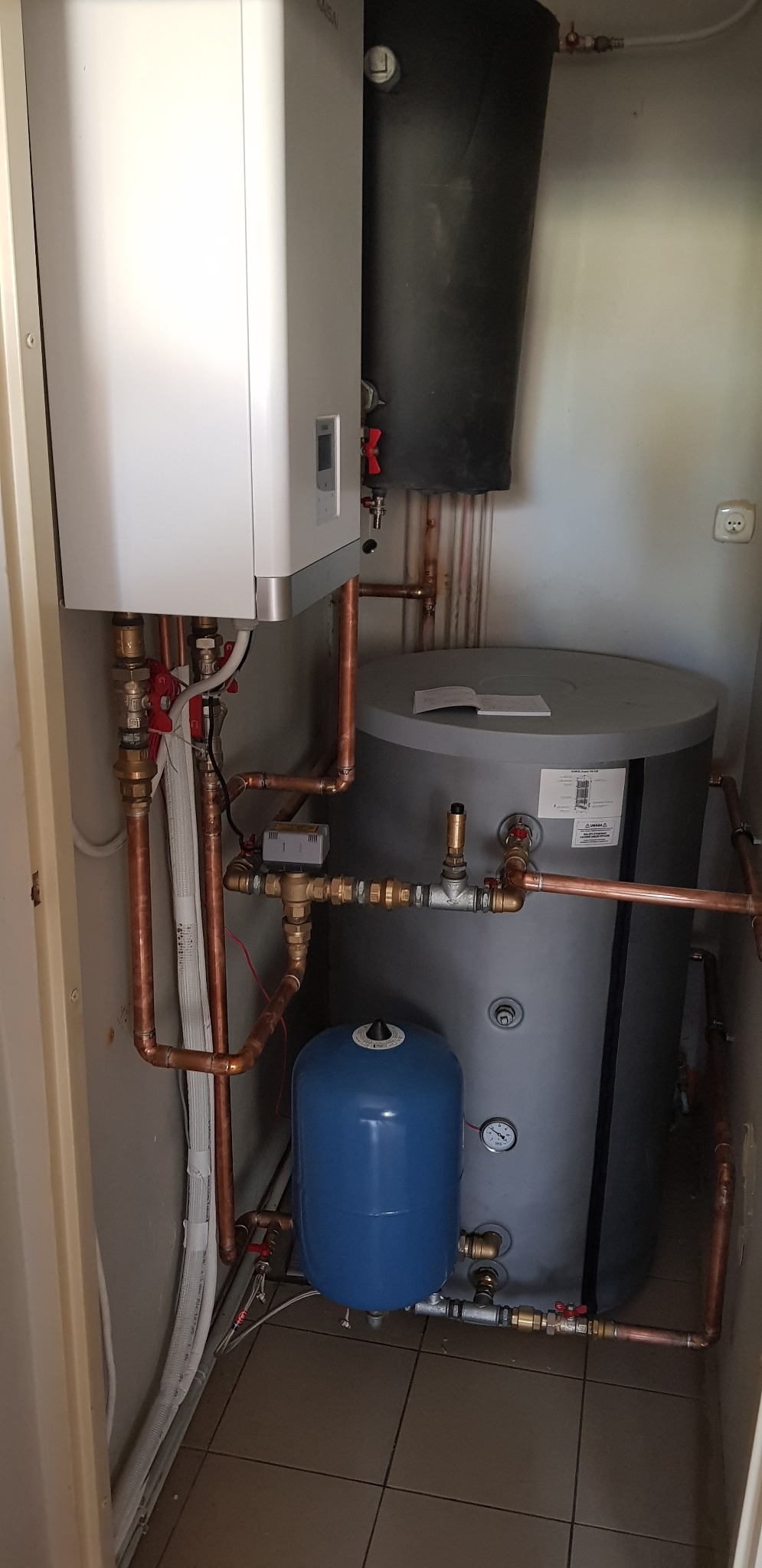 Zestaw urządzeń grzewczych w kotłowni: biały piec, szary zasobnik ciepłej wody, czarny zbiornik buforowy i niebieskie naczynie przeponowe, połączone miedzianymi rurami.