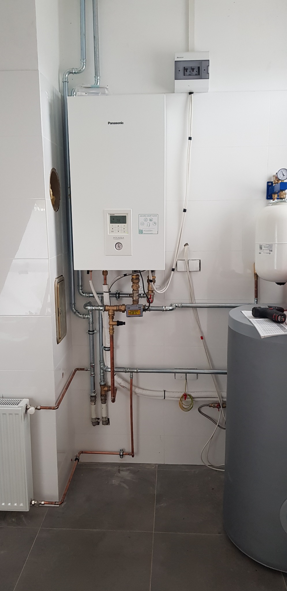 Biały piec grzewczy Panasonic z widocznymi rurami miedzianymi i elementami instalacji w pomieszczeniu z białymi płytkami, obok szary zbiornik i narzędzie na górze.