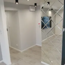 Nowoczesny salon z szarą podłogą z płytek, lustrzaną ścianą z romboidalnych paneli, białymi ścianami i czarnymi lampami sufitowymi