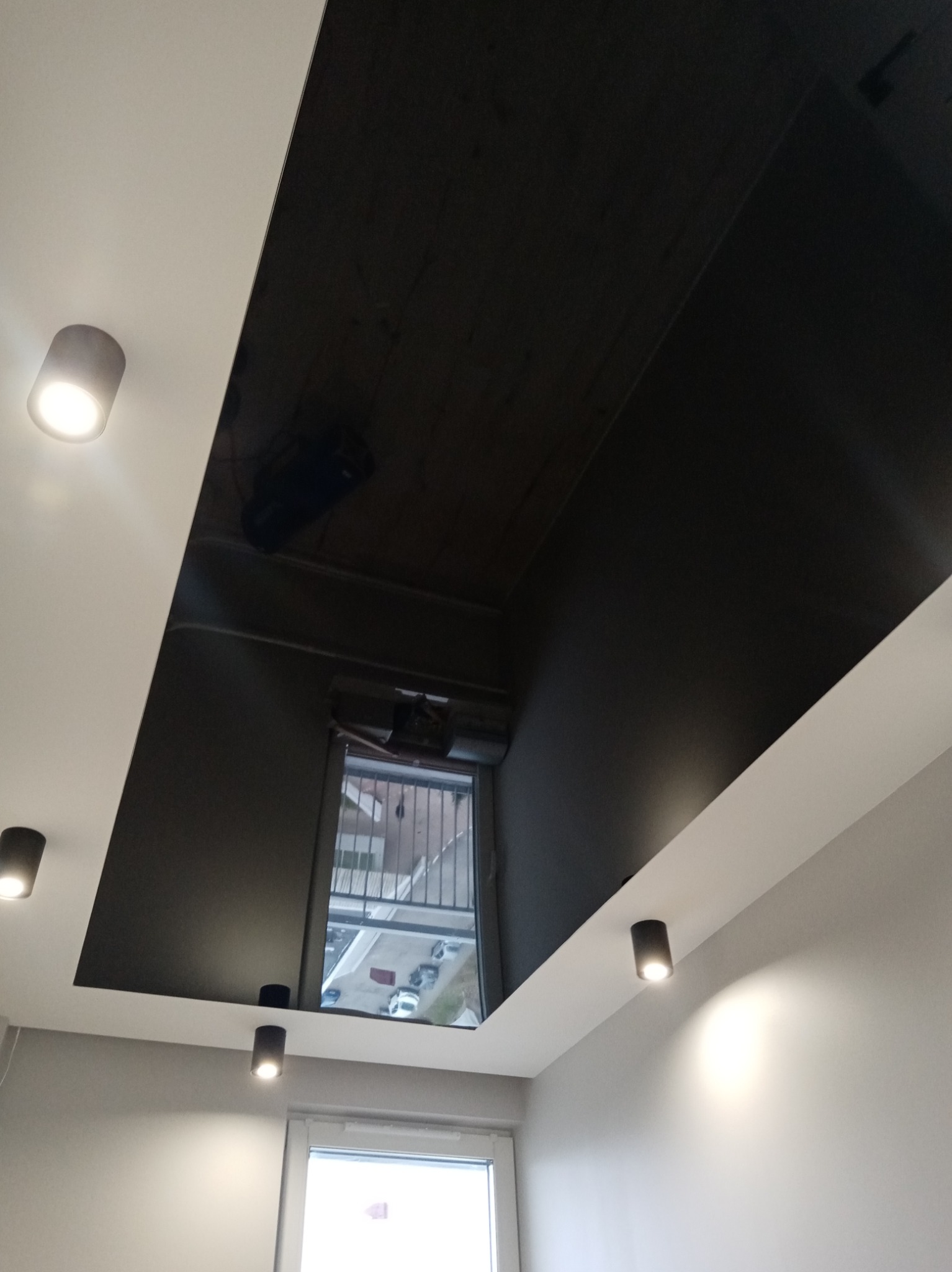 Część sufitu z czarną, błyszczącą wstawką, w której odbija się okno, otoczona białym sufitem z zamontowanymi punktowymi lampami LED.
