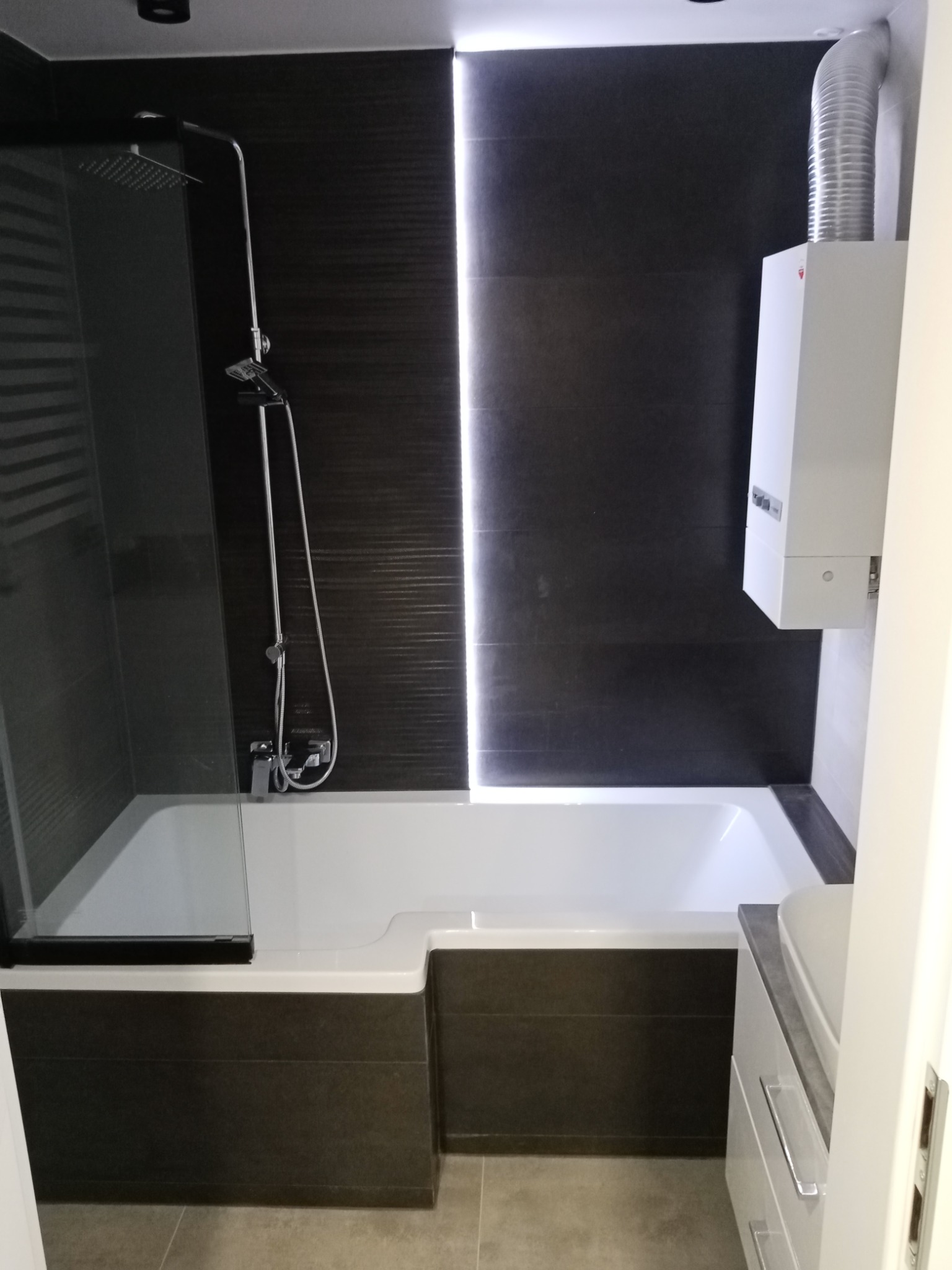 Nowoczesna łazienka z kabiną prysznicową nad wanną, ciemnymi płytkami na ścianie i białym grzejnikiem z rurą wentylacyjną.